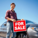 A person stands next to a well-maintained car, holding a "For Sale" sign on a sunny day with a clear blue sky, conveying positivity and opportunity.